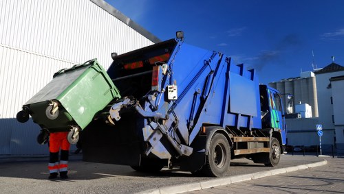 A large blue and black commercial waste collection vehicle is positioned on a paved street, actively lifting a green wheeled dumpster with its hydraulic arm. The green dumpster, made of plastic with visible textured surface, is tilted at an angle as it is emptied into the vehicle's open hopper. The waste collection vehicle has a prominent blue body with black chassis, distinct hydraulic components, and safety reflectors on the back. In the background, there is a white corrugated metal building to the left and a clear blue sky with a few clouds. A person wearing a high-visibility safety vest and red trousers stands nearby, overseeing the operation. The scene appears to be set in an urban or commercial area, possibly within the postcode area associated with Maida Vale, west London, consistent with the context of commercial waste collection services provided by Commercial Waste Maida Vale.