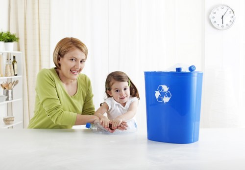A woman with short blonde hair and wearing a light green top is seated at a white table, smiling while looking at the camera. Next to her, a young girl with brown hair in pigtails, wearing a white top, also faces the camera with a slight smile. On the table in front of them is a large blue plastic recycling bin with a white recycling symbol printed on its front, positioned on a clean, bright surface. The background features a white wall with a simple clock showing the time as approximately 12:02, and a window with light-colored curtains allowing natural light to fill the scene. The environment appears to be a domestic setting, possibly a kitchen or utility area, emphasizing a focus on recycling and waste management, consistent with services offered by companies such as Commercial Waste Maida Vale in the local postcode vicinity of Maida Vale, London, for rubbish collection and waste disposal.