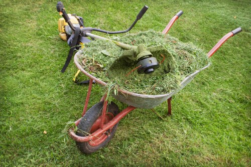 A red wheelbarrow filled with freshly cut grass and garden debris, including patches of green foliage and small twigs, situated on a well-maintained grassy lawn. The wheelbarrow has a black rubber tyre with visible treads and metal handles at the rear, which extend outward and slightly upward. Behind the wheelbarrow, a garden trimmer with a black handlebar, yellow engine casing, and metallic shaft rests on the grass, indicating active yard maintenance. The surrounding environment features lush, green grass with a uniform cut, typical of residential or urban garden settings, possibly in Maida Vale. The scene is evenly lit, likely during daylight hours, emphasizing the textures of the grass, the grass clippings, and the metallic and plastic surfaces of the equipment. This image illustrates a typical gardening or waste clearance activity, aligning with the services provided by Commercial Waste Maida Vale for garden waste removal and general rubbish clearance.