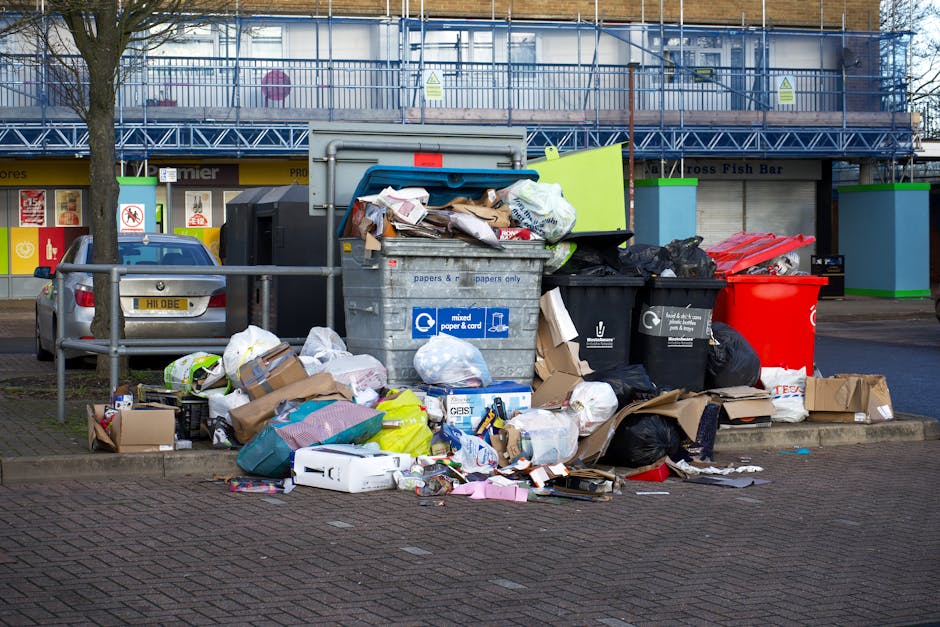 A small, weathered blue pickup truck parked on the side of a paved street during daylight hours, loaded with a large amount of mixed waste, including cardboard boxes, black plastic bags, and various o