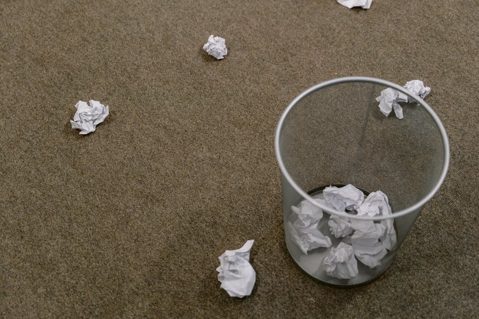 A clear cylindrical waste bin made of transparent plastic with a metal rim at the top is positioned on a brown textured carpeted floor. Inside the bin, there are crumpled sheets of white paper, some o