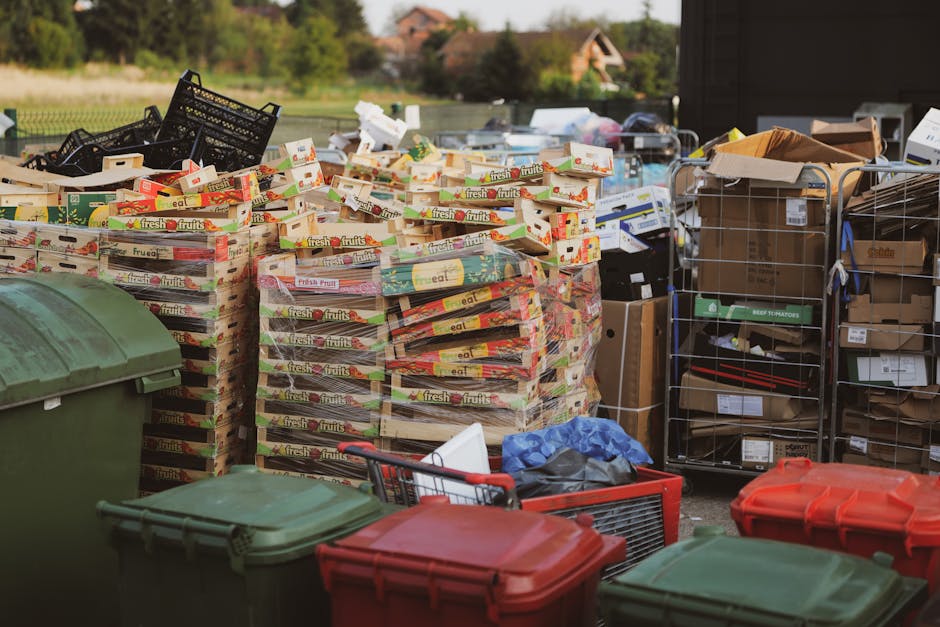 A row of large, wheeled rubbish bins lined up outdoors beside a brick wall, with the front green bin distinctly visible and other blue, red, and green bins extending into the background. The green bin