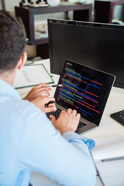 A person wearing a light blue shirt is sitting at a desk and working on a silver laptop that displays lines of colorful computer code on its screen, with a large black monitor positioned behind it. Th